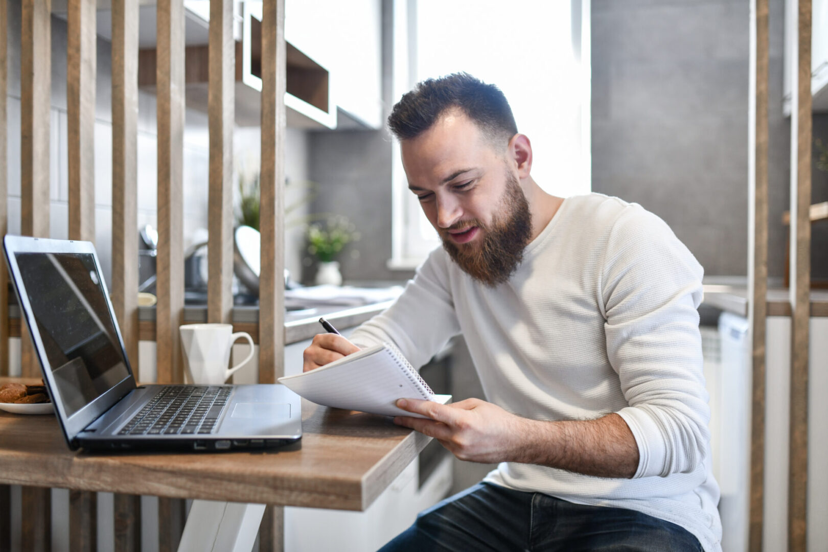Man writing notes at laptop in kitchen.
